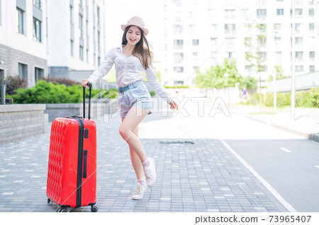 Young caucasian woman in a hat and shorts stands on the street with a red suitcase. Young caucasian woman in a hat and shorts stands on the street with a red suitcase. 73965407