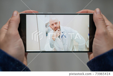 Closeup of female hands with a phone. A bald man doctor remotely consults a patient with a webcam. The woman says with a friendly physician for video calls. 73965414