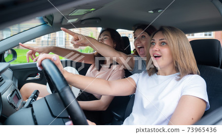 Three friends go on a trip. Caucasian women drive in a car and point fingers in surprise. 73965436