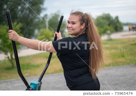 Beautiful smiling overweight young woman does fitness on an ellipsoid simulator outdoors. Fat girl is training on the sports ground for weight loss. Sport outside on a warm summer day. Beautiful smiling overweight young woman does fitness on an ellipsoid simulator outdoors. Fat girl is training on the sports ground for weight loss. Sport outside on a warm summer day. 73965474