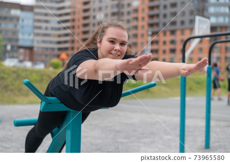 Fat young woman doing back extension exercise on exercise machines on the sports ground outdoors. Fat young woman doing back extension exercise on exercise machines on the sports ground outdoors. 73965580