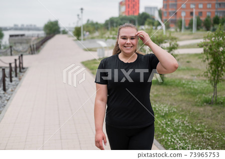 An obese young woman jogging outdoors. Fat beautiful smiling girl in a black tracksuit is engaged in fitness for weight loss on the waterfront. A woman runs on a summer day. 73965753