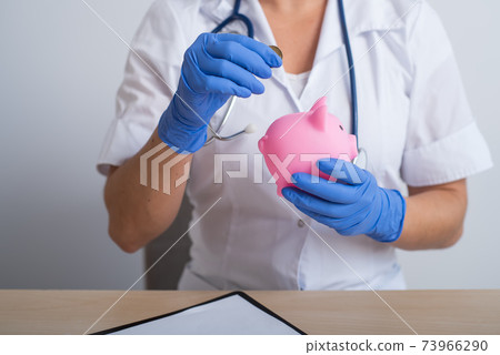 A faceless female doctor puts a coin in a piggy bank 73966290