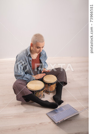 A young hipster woman sits on the floor at home and watches educational videos on mini bongs. Girl remotely learns to play traditional ethnic drums. A young hipster woman sits on the floor at home and watches educational videos on mini bongs. Girl remotely learns to play traditional ethnic drums. 73966581