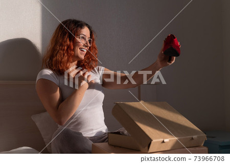 Joyful woman opens ordered shoes while sitting in bed. Online shopping concept with home delivery Joyful woman opens ordered shoes while sitting in bed. Online shopping concept with home delivery 73966708