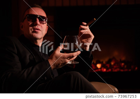 A mature man with glasses drinks brandy and smokes a cigar while sitting in an armchair by the fireplace in the dark. The concept of an elite gentlemen's club A mature man with glasses drinks brandy and smokes a cigar while sitting in an armchair by the fireplace in the dark. The concept of an elite gentlemen's club 73967253