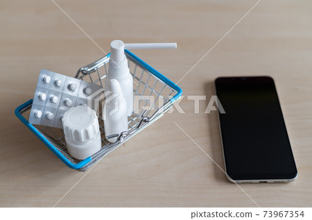 Online pharmacy. Blister with pills throat spray jar with capsules in a mini shopping basket next to a mobile phone with a black screen. Smartphone app for buying medicine. 73967354