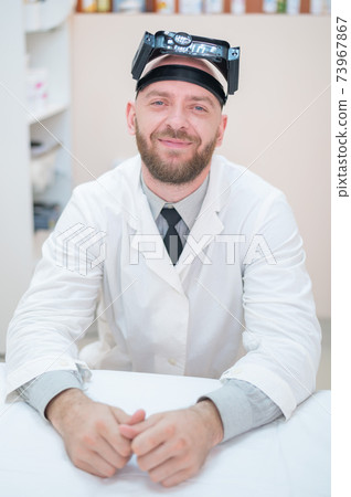 Male bearded doctor in a medical coat and with a magnifying head strap w lights sitting on the desk. Optometrist equipment. Medical office. Male bearded doctor in a medical coat and with a magnifying head strap w lights sitting on the desk. Optometrist equipment. Medical office. 73967867