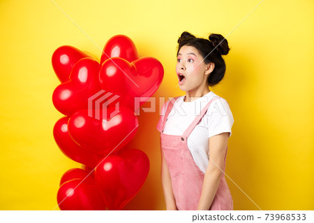 Valentines day and relationship concept. Fashion girl looking amazed, gasping and say wow, staring left while standing near red hearts balloons, surprise from lover, yellow background 73968533