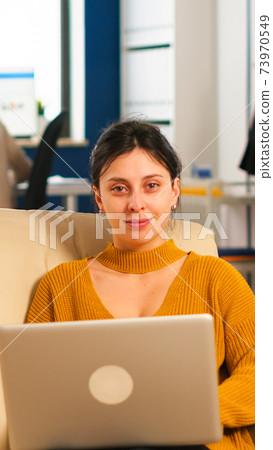 Businesswoman sitting on couch holding laptop, smiling at camera 73970549