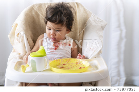 Baby adorable girl with apron holding spoon in her hand while sitting on the chair eating cake  73977055