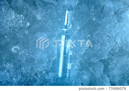 vaccine in glass transparent ampoule with clear liquid with medical preparation for treatment of viral disease is cool to subzero temperatures lying on frozen ice, blue background on pharmaceutical. 73986079