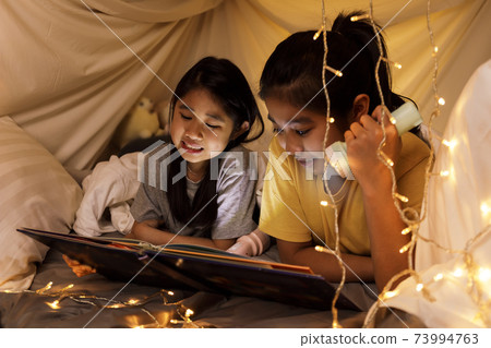 Family concept. elder sister and sister reading book with flashlight together in children tent before bedtime. Happy elder sister read story book to her sister in bed sheet tent. 73994763
