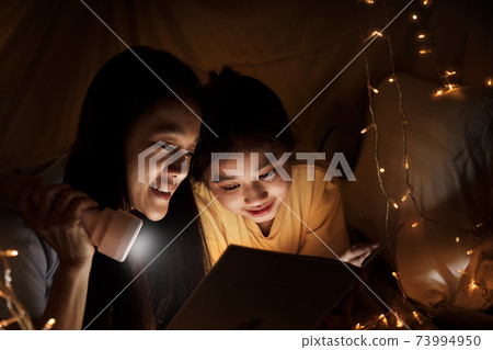 Family concept. elder sister and sister reading book with flashlight together before bedtime. Sister read story book together in bed sheet tent. focus selective little sister. With film grain effect 73994950