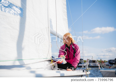 A purposeful young female yachtsman pulls the rigging rope to raise the sail on the yacht. 73996796