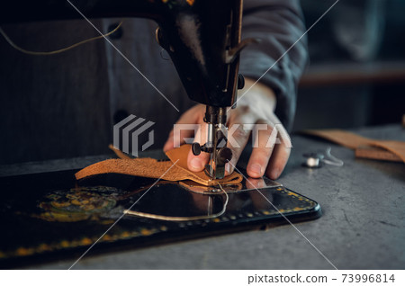 Close-up photo of a high-quality thread seam on a piece of brown genuine leather. Hands of a tailor and a sewing machine 73996814