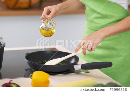 Young woman housewife cooking in the kitchen while adding olive oil. Concept of fresh and healthy meal at home Young woman housewife cooking in the kitchen while adding olive oil. Concept of fresh and healthy meal at home 73996818