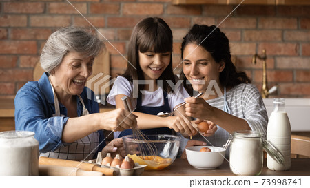 Happy little girl with mother and grandmother cooking, stirring eggs Happy little girl with mother and grandmother cooking, stirring eggs 73998741