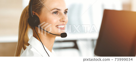 Blond female customer service representative is consulting clients online using headset in sunny office. Call center concept Blond female customer service representative is consulting clients online using headset in sunny office. Call center concept 73999034