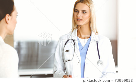 Woman-doctor at work in sunny hospital is happy to consult female patient. Blonde physician checks medical history record and exam results while using clipboard Woman-doctor at work in sunny hospital is happy to consult female patient. Blonde physician checks medical history record and exam results while using clipboard 73999038
