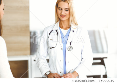 Woman-doctor at work in sunny hospital is happy to consult female patient. Blonde physician checks medical history record and exam results while using clipboard Woman-doctor at work in sunny hospital is happy to consult female patient. Blonde physician checks medical history record and exam results while using clipboard 73999039