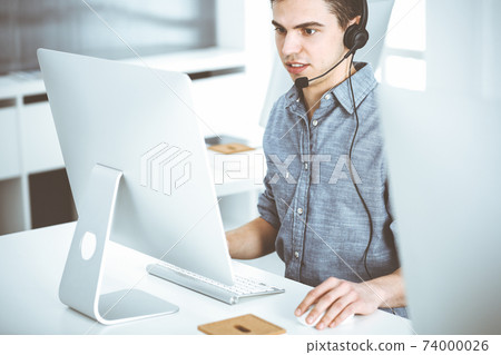 Casual dressed young man using headset and computer while talking with customers online. Call center, business concept Casual dressed young man using headset and computer while talking with customers online. Call center, business concept 74000026