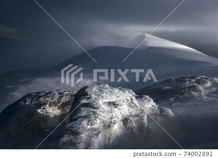 Snow covered winter mountains  in last evening sunlight. Magnificent windy dusk on tops above picturesque alpine ski resort 74002891