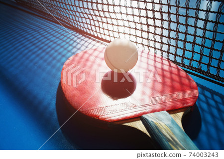 A red table tennis racket and a white ball lie on the surface of the table next to the net. Sports A red table tennis racket and a white ball lie on the surface of the table next to the net. Sports 74003243