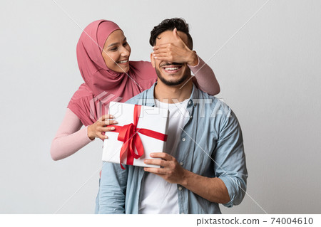Loving muslim woman in hijab surprising her husband with gift box Loving muslim woman in hijab surprising her husband with gift box 74004610