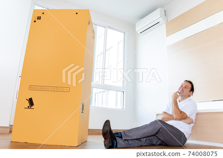 Man sitting on the floor in an empty apartment looking at a big cardboard box.  74008075