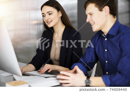 Young business people are talking to each other, while sitting at the desk in sunny office. Businesswoman is explaining something to her colleague Young business people are talking to each other, while sitting at the desk in sunny office. Businesswoman is explaining something to her colleague 74009440