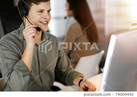 Young dark-haired guy with headsets is talking to a client, while sitting in sunny office. Call center operators at work Young dark-haired guy with headsets is talking to a client, while sitting in sunny office. Call center operators at work 74009442