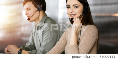 Call center operators at work. Two young people in headsets are talking to the clients, while sitting in sunny office Call center operators at work. Two young people in headsets are talking to the clients, while sitting in sunny office 74009443