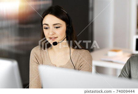 Young friendly girl in headsets is talking to a firm's client, while sitting at the desk in sunny office. Call center operators at work Young friendly girl in headsets is talking to a firm's client, while sitting at the desk in sunny office. Call center operators at work 74009444