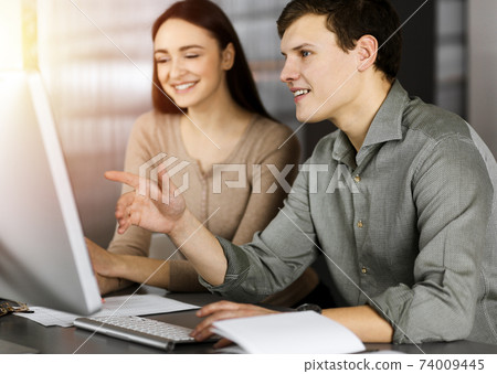 Friendly young businessman in a green shirt is working on computer, while sitting together at the desk with a female colleague in a modern office Friendly young businessman in a green shirt is working on computer, while sitting together at the desk with a female colleague in a modern office 74009445