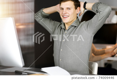 Young businessman with brown curly hair in a green shirt is stretching his hands after working hard on computer in sunny office Young businessman with brown curly hair in a green shirt is stretching his hands after working hard on computer in sunny office 74009446