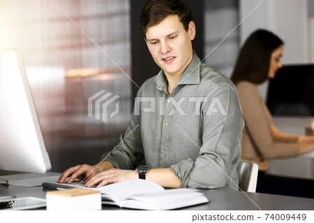 Young businessman working on his computer sitting at the desk in office. Headshot portrait of a man Young businessman working on his computer sitting at the desk in office. Headshot portrait of a man 74009449