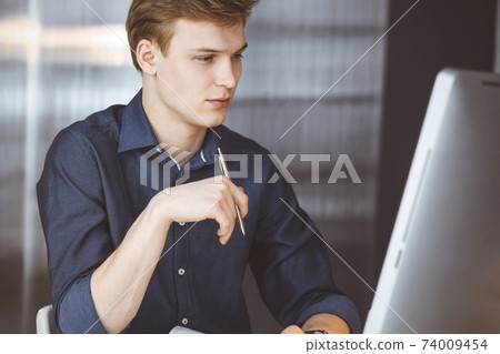 Young blond businessman thinking about strategy at his working place with computer. Startup business means working hard and out of time for success achievement Young blond businessman thinking about strategy at his working place with computer. Startup business means working hard and out of time for success achievement 74009454