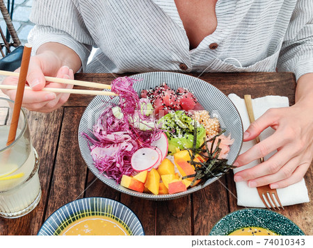 Woman eating tasty colorful healthy natural organic vegetarian Hawaiian poke bowl using asian chopsticks on rustic wooden table. Healthy natural organic eating concept Woman eating tasty colorful healthy natural organic vegetarian Hawaiian poke bowl using asian chopsticks on rustic wooden table. Healthy natural organic eating concept 74010343