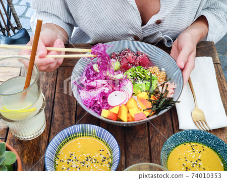 Woman eating tasty colorful healthy natural organic vegetarian Hawaiian poke bowl using asian chopsticks on rustic wooden table. Healthy natural organic eating concept Woman eating tasty colorful healthy natural organic vegetarian Hawaiian poke bowl using asian chopsticks on rustic wooden table. Healthy natural organic eating concept 74010353