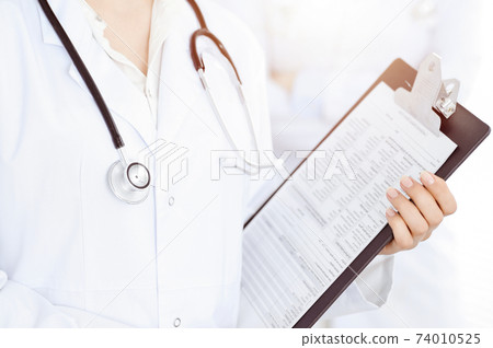 Unknown woman-doctor standing straight with clipboard of medication history record in clinic, close-up. Medicine concept Unknown woman-doctor standing straight with clipboard of medication history record in clinic, close-up. Medicine concept 74010525