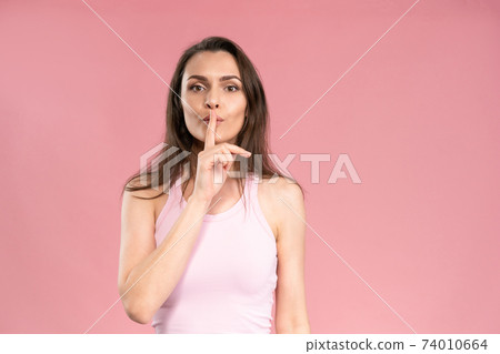 Gesture with a finger to the mouth says to be quiet or keep a secret young woman with dark hair in trendy pink t-shirt looking at camera. Facial expressions, emotions, feelings 74010664