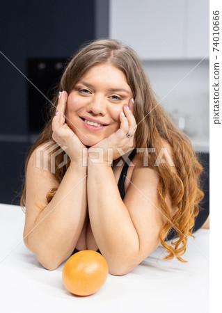 Girl with obese problem. Young sexy chubby white girl in black swimsuit, white shirt at modern kitchen table. juggling fruits in hand. Trying to loose weight fast. Fat barefoot girl low carb diet 74010666