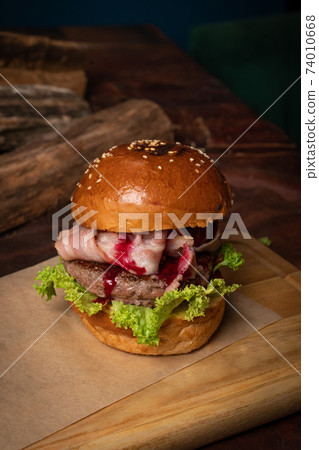 Craft burger with cherry sauce, fried slices of bacon, fresh lettuce next to the decorative wooden lodges. Dish served on a wooden cutting board and wood on background. Street food restaurant concept 74010668