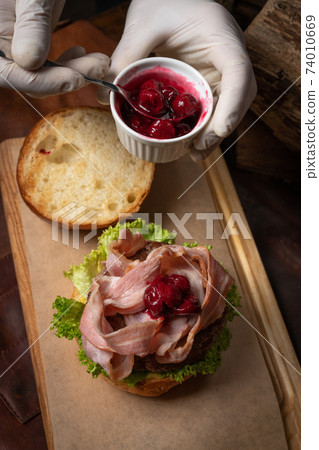 Top view. Chef serving fresh craft beef burger with fried slices of bacon covering with sweet and sour cherry sauce and fresh lettuce before serving it to a client. Street food concept 74010669