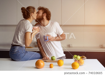 Kissing beautiful couple embracing each other enjoying their their morning coffee in a new house. Making fun at modern kitchen and kissing while cooking at home. New house concept Kissing beautiful couple embracing each other enjoying their their morning coffee in a new house. Making fun at modern kitchen and kissing while cooking at home. New house concept 74010685