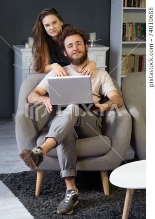 Working together young couple staying quarantined at home with sitting on the arm chairs. Happy young couple sitting on a chair together using laptop. Square cropped Working together young couple staying quarantined at home with sitting on the arm chairs. Happy young couple sitting on a chair together using laptop. Square cropped 74010688