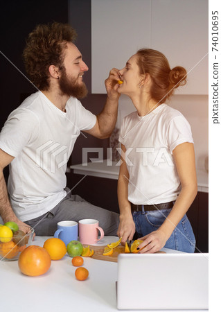 Feeding orange to girl young man embracing each other enjoying their their morning coffee in a new house. Making fun at modern kitchen and kissing while cooking at home. New house concept Feeding orange to girl young man embracing each other enjoying their their morning coffee in a new house. Making fun at modern kitchen and kissing while cooking at home. New house concept 74010695