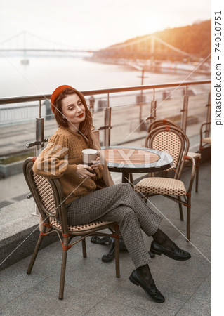 Charming woman enjoying her cup of coffee sitting in a spring outdoor cafe. French woman in red beret with background of urban city. Parisian woman with coffee mug on city street in evening Charming woman enjoying her cup of coffee sitting in a spring outdoor cafe. French woman in red beret with background of urban city. Parisian woman with coffee mug on city street in evening 74010751