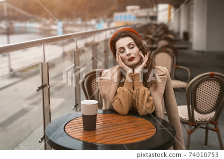 Parisian woman with coffee mug on city street in evening. Charming woman enjoying her cup of coffee sitting in a spring outdoor cafe. French woman in red beret with background of urban city Parisian woman with coffee mug on city street in evening. Charming woman enjoying her cup of coffee sitting in a spring outdoor cafe. French woman in red beret with background of urban city 74010753